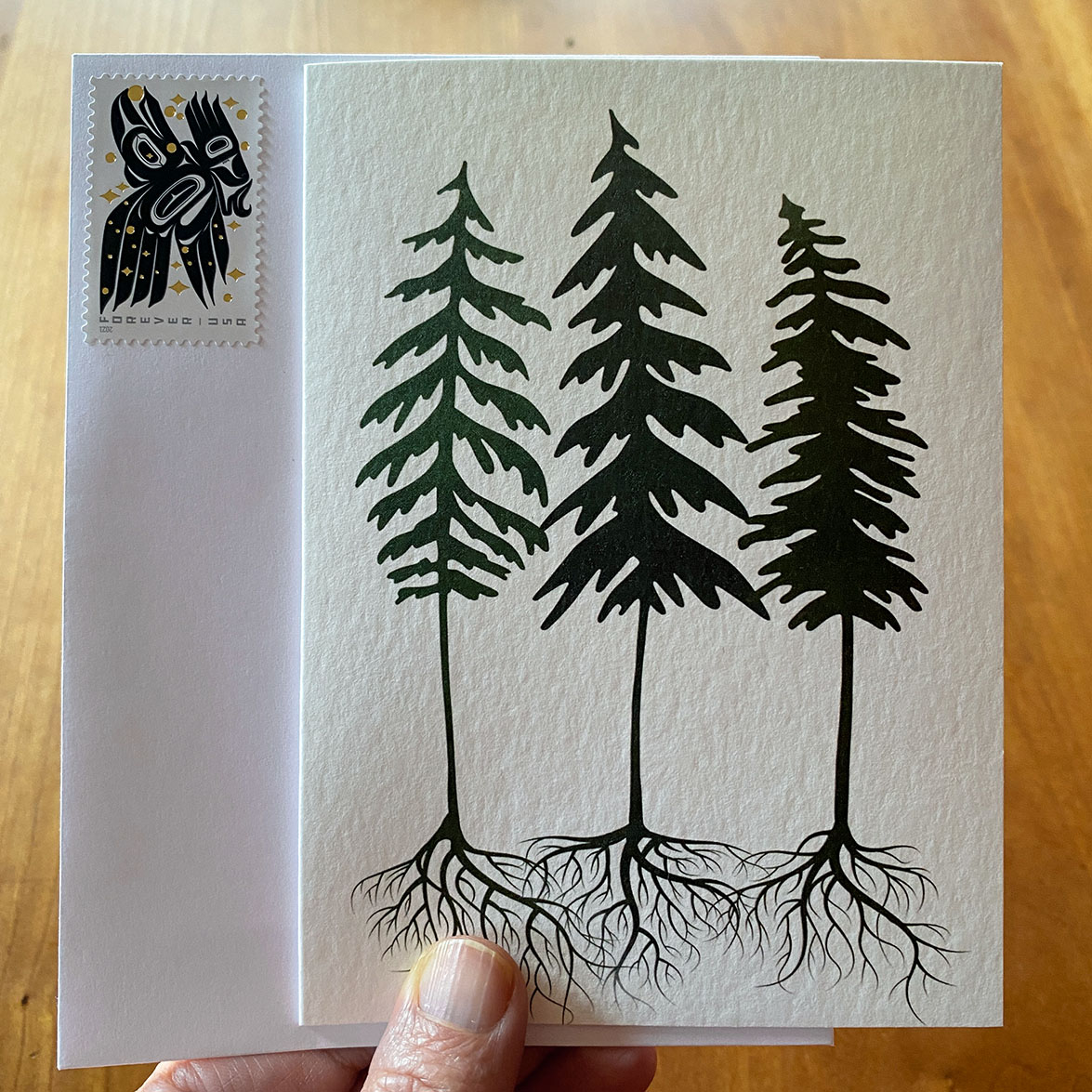 Three fir trees stand together with roots entwined on a gratitude card with envelope and raven stamp.