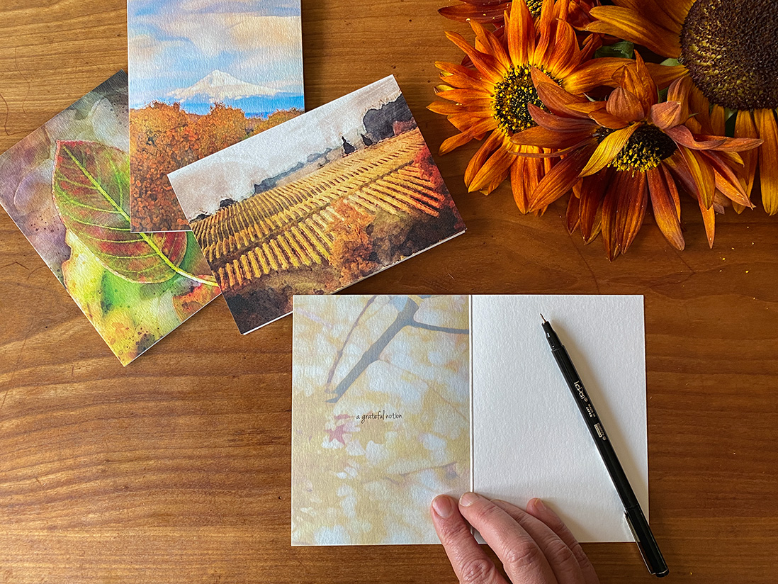 Three fall-themed gratitude cards from Kindness Roots with a hand about to write on them with a black pen and sunflowers on the table near by.